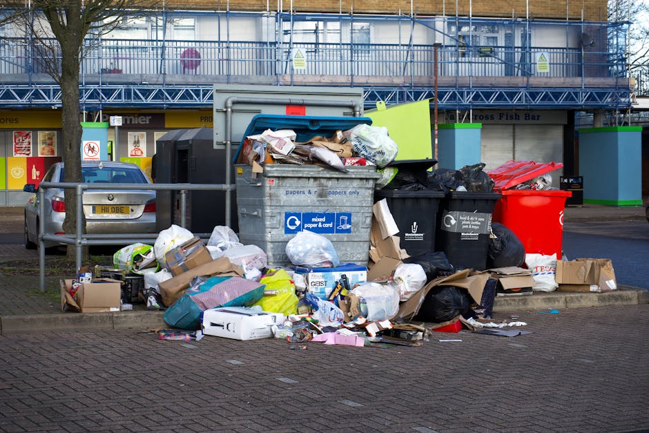 A collection of overflowing waste and rubbish bags positioned on a paved sidewalk in front of a small parking area, with a metal railing separating the waste from a silver car parked nearby. The waste includes large black garbage bags, flattened cardboard boxes, and assorted paper and packaging materials scattered around. In the background, there are commercial storefronts with signs and a blue metal construction barrier or scaffolding covering part of a building. A grey wheelie bin with a blue lid is visibly stacked with uncollected rubbish, and other recycling bins in black and red are present nearby, partially filled with additional trash. The scene appears to be an outdoor area designated for waste disposal, highlighting the need for effective rubbish removal services, such as those provided by Waste Collection Harringay, especially in contexts of private waste collection or on-site clearance, with natural daylight illuminating the area and emphasizing the uncollected rubbish's volume and disorganization.