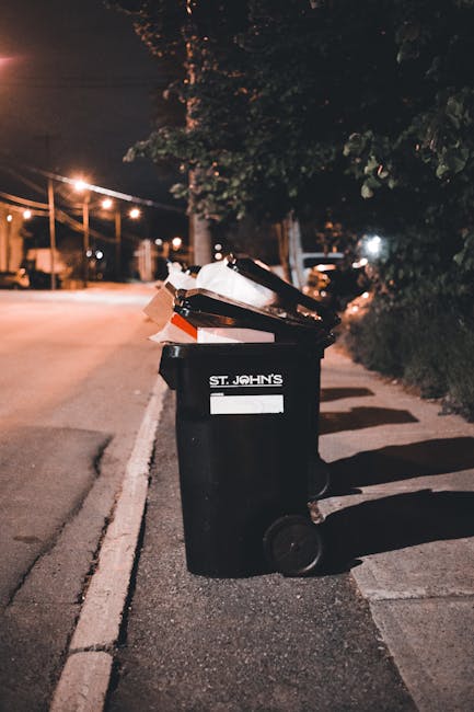 A black wheeled rubbish bin with a white label and the name 'St. John's' printed on it, positioned on a paved sidewalk beside a street at night. The bin is filled with various discarded items, including cardboard boxes and plastic wrappers, which are loosely stacked and protruding from the open lid. The surrounding environment features a dimly lit residential area with streetlights casting a warm glow, tall trees with leafy branches partially illuminated, and parked vehicles visible in the background. The scene suggests an outdoor waste collection point, highlighting the use of private rubbish disposal services such as those offered by Waste Collection Harringay, especially during evening hours when street-side bins are often emptied or awaiting collection. The shadows cast by the bin and nearby objects add depth to the setting, creating a quiet, urban atmosphere focused on domestic waste management.