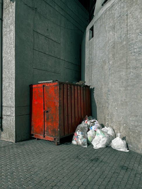 A collection of overflowing waste and rubbish bags positioned on a paved sidewalk in front of a small parking area, with a metal railing separating the waste from a silver car parked nearby. The waste includes large black garbage bags, flattened cardboard boxes, and assorted paper and packaging materials scattered around. In the background, there are commercial storefronts with signs and a blue metal construction barrier or scaffolding covering part of a building. A grey wheelie bin with a blue lid is visibly stacked with uncollected rubbish, and other recycling bins in black and red are present nearby, partially filled with additional trash. The scene appears to be an outdoor area designated for waste disposal, highlighting the need for effective rubbish removal services, such as those provided by Waste Collection Harringay, especially in contexts of private waste collection or on-site clearance, with natural daylight illuminating the area and emphasizing the uncollected rubbish's volume and disorganization.
