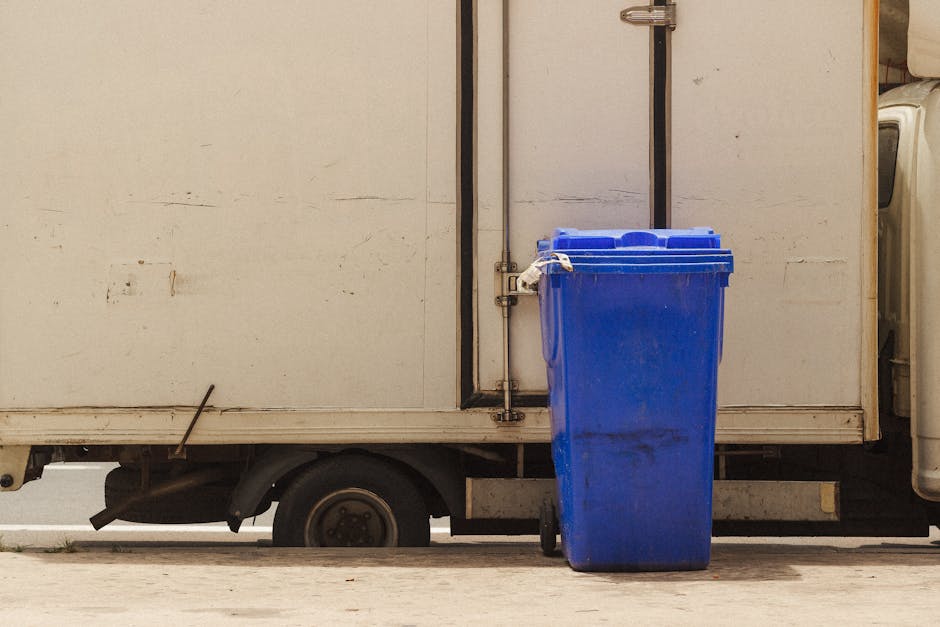 A blue wheeled rubbish bin made of plastic with a textured surface and a slightly dirty appearance, positioned adjacent to a white delivery or storage truck parked on a paved surface. The bin is upright, with the lid closed, and positioned near the rear side of the truck, which features a plain, slightly weathered exterior with visible scratches and dirt marks. The truck's rear wheel and part of the chassis are visible beneath the cargo area. The environment appears to be an outdoor loading or collection point, with diffused natural lighting suggesting daytime. The scene relates to waste collection, highlighting the use of private waste disposal methods such as bin transfer and on-site collection, with Waste Collection Harringay potentially involved in managing such rubbish removal activities. The focus remains on the practical aspect of rubbish handling with an emphasis on waste container placement and access in a professional context.