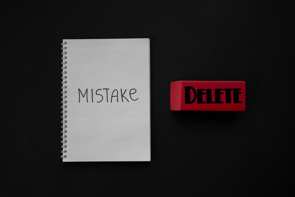 A white spiral-bound notebook with the word 'MISTAKE' written in black capital letters on the cover, positioned on a plain black background. To the right of the notebook, there is a red rubber stamp with the word 'DELETE' in bold black letters, lying flat on the surface. The image conveys a concept of correcting errors or removing unwanted items, which may relate to proper waste handling or avoiding disposal mistakes in the context of rubbish removal services. The lighting is even, without reflections or shadows, emphasizing the stark contrast between the white notebook, red stamp, and black background, aligning with a professional and neutral tone suitable for waste management topics or alternative waste collection considerations.