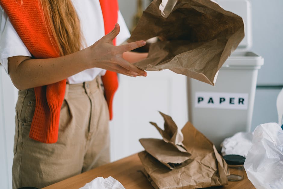 Close-up of a person wearing a white shirt and an orange vest, holding an empty brown paper bag over a cluttered table in an indoor setting. The table has crumpled paper and several other partially opened paper bags, indicating a waste or rubbish collection process. In the background, a white waste bin labeled 'PAPER' is visible, suggesting an organized system for segregating waste materials, typical of private or alternative disposal methods. The environment appears well-lit with natural or fluorescent lighting, emphasizing the texture and color of the paper bags, which are made of kraft paper with rough, fibrous finishes. The person’s hands, with visible skin tones and clean fingernails, are actively engaged in lifting or stacking waste items, highlighting a process related to rubbish removal or household waste sorting. This scene subtly reflects the importance of organized waste management, aligning with practices like on-site clearance or independent collection services offered by companies such as Waste Collection Harringay, in accordance with local rubbish rules and regulations.
