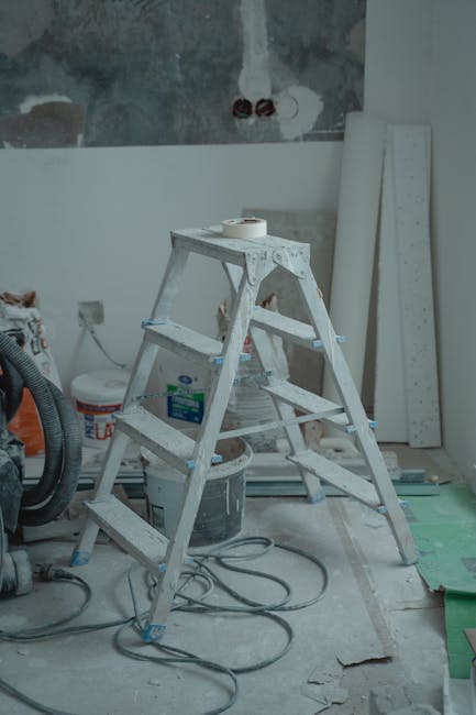 The image shows an indoor construction or renovation scene with a white, multi-step metal platform ladder positioned centrally. The ladder has visible paint splatters and is placed on a concrete or unfinished floor. Surrounding the ladder are various materials and tools, including a large plastic bucket, a roll of masking tape on top of the ladder, and an insulated cable lying on the floor. To the right, there are long, white rectangular panels or boards leaning against the wall, which has an unfinished, textured appearance with patches of plaster and exposed brickwork. In the background, an electrical outlet and some wiring are visible on the wall, indicating ongoing electrical work. The lighting appears natural or from overhead sources, casting soft shadows across the scene. This setting suggests a workspace prepared for or involved in on-site renovation or repair activities, where waste and debris are likely to be generated, aligning with the services provided by waste collection specialists such as Waste Collection Harringay, who handle rubbish removal during property refurbishments.