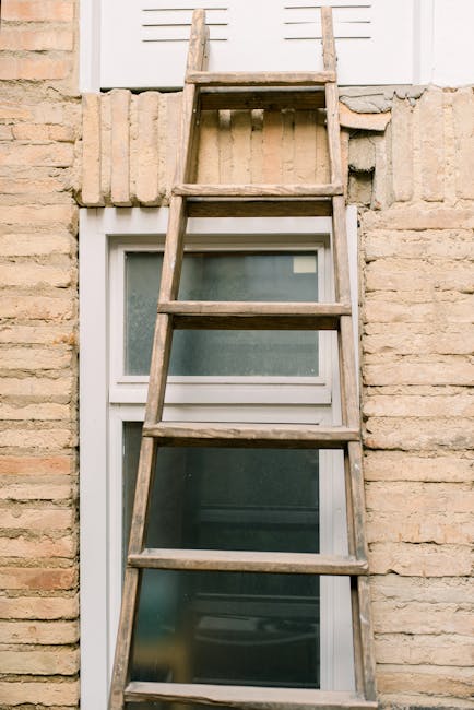 A wooden ladder with four rungs leaning against the exterior of a brick building, positioned above a double-glazed window with a white frame. The ladder appears weathered, with visible natural wood grain and slight discoloration, and extends from ground level up to a white window shutter or panel. The brick wall is composed of beige and light brown bricks with a coarse texture, arranged in a running bond pattern, with some mortar joints visible. The window below has frosted or semi-opaque glass, with a slight reflection or tint, set within a white PVC frame that contrasts with the brickwork. The environment suggests an outdoor wall of a residential or commercial property, with the ladder possibly used for access or maintenance, indicating a scenario of private on-site clearance or repairs related to waste or property upkeep. The lighting appears natural, with soft shadows suggesting daylight conditions. Waste collection or clearance services such as those from Waste Collection Harringay could handle debris or materials associated with such exterior maintenance work.