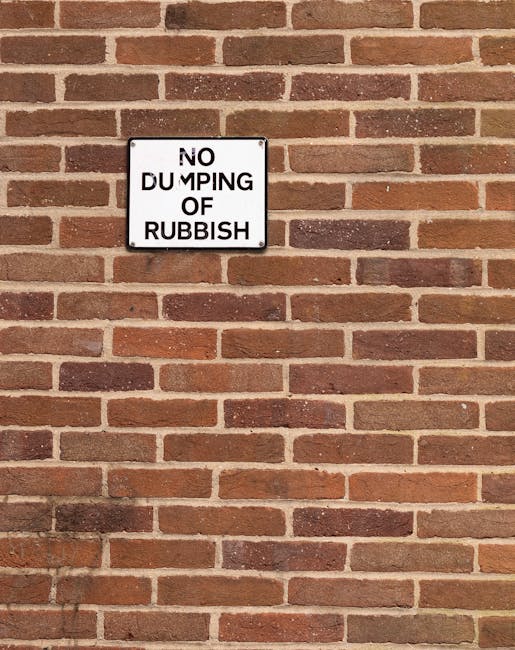 A rectangular metal sign mounted on a red and brown brick wall, displaying the message 'NO DUMPING OF RUBBISH' in black uppercase letters with a white background and a black border. The bricks have a textured surface with variations in colour, arranged in a horizontal pattern with mortar joints in between. The sign is positioned centrally near the upper left section of the wall, which appears to be part of a building exterior. The lighting is natural, casting soft shadows beneath the sign. This scene relates to regulations around waste disposal and highlights the importance of proper rubbish removal, consistent with services offered by waste collection providers such as Waste Collection Harringay, particularly in the context of local or private rubbish management near residential or commercial premises.