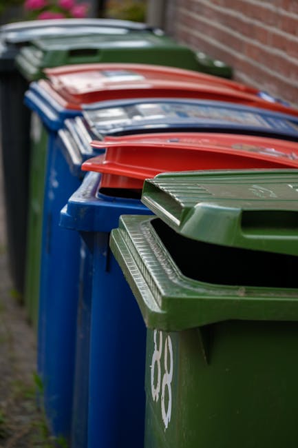 A row of large, rectangular wheelie bins positioned along a pavement, each with a distinct color and fitted with a hinged lid. The first bin in the foreground is green with a textured lid, followed by a blue bin, and then lids in red, blue, and green extending into the background. The bins are made of durable plastic with raised ridges on the lids for grip. They are placed close together, aligned along a curb beside a brick wall, with a narrow paved walkway visible on the left. The environment appears to be an urban residential area, with natural daylight illuminating the scene, emphasizing the different colors and textures of the bins. The overall scene suggests arrangements for waste collection and disposal, indicative of a private or alternative rubbish removal service, like those offered by Waste Collection Harringay.