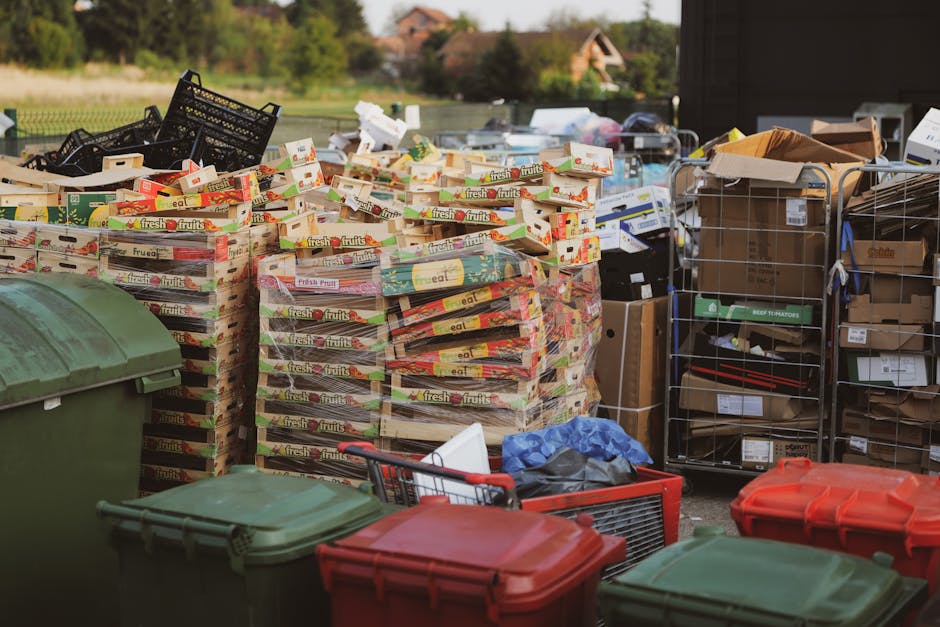 An outdoor scene featuring a large pile of stacked cardboard boxes with colorful fruit juice labels, primarily yellow, red, and green, arranged on wooden pallets. To the right, there are metal wire cages filled with various packaging materials, including cardboard boxes, plastic containers, and smaller assorted items. In the foreground, several large green and red wheelie bins with closed lids are positioned on a paved area, and a small shopping trolley containing a black plastic bag and a piece of white foam. Behind the containers, a background of lush greenery and trees is visible, along with a glimpse of distant brick houses under a clear sky, indicating a residential or semi-rural setting. The scene appears to depict a private waste collection or on-site clearance, possibly arranged by waste management services like Waste Collection Harringay, with an emphasis on waste that includes cardboard, plastics, and packaging materials, relevant to non-urban waste disposal or alternative rubbish removal options.