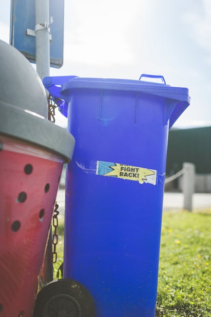 A row of large, wheeled rubbish bins lined up along a pavement outside a brick building, with green, blue, and red lids. The bins are made of durable plastic, with textured lids and smooth sides, and are situated close together, extending from the foreground to the background. The green bin at the front is marked with a white recycling symbol, indicating its use for recyclable waste, while the other bins are plain. The scene shows natural daylight with a slightly overcast sky, casting soft shadows on the bins. The setting suggests an outdoor waste collection point, typical for communal or private rubbish removal services, such as those offered by Waste Collection Harringay, which specialises in alternative waste handling and on-site clearance solutions.