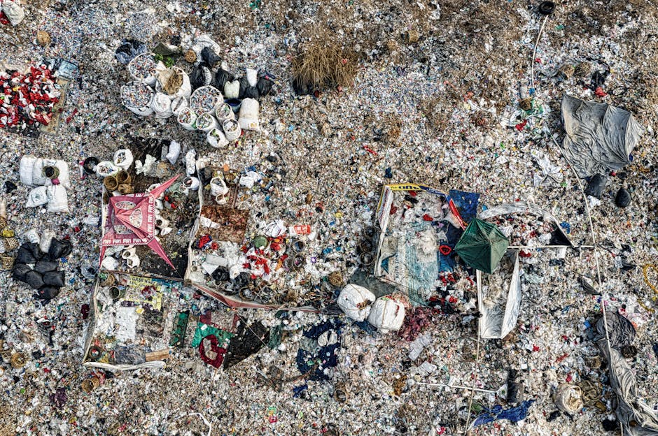 A vast expanse of littered ground covered with a mixture of waste materials including crushed plastic bottles, aluminum cans, torn plastic bags, cardboard fragments, and paper debris. The trash is scattered unevenly across the gravelly surface, with some items piled together and others strewn loosely. Several black and white plastic bags are crumpled and dispersed among the debris, while aluminum cans are flattened and mixed with food wrappers and paper packaging. A partially collapsed pink and white canopy tent with metal poles lies amidst the rubbish, alongside a green and red fabric item that appears to be part of the tent or a similar structure. The surrounding environment appears to be an outdoor area, possibly a landfill or illegal dumping site, with no visible vegetation or natural features. The scene is lit with natural daylight, casting minimal shadows, highlighting the texture and chaos of the discarded waste, which reflects a typical scenario of unmanaged refuse and the need for rubbish collection services such as those offered by Waste Collection Harringay.