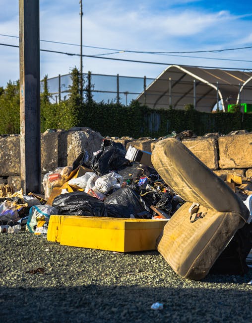 A close-up view of a roadside rubbish pile consisting of black plastic trash bags, discarded packaging, and miscellaneous waste materials, situated on a paved surface next to a stone wall. The waste is spilling out of a yellow container and appears irregularly stacked, with some items leaning against an old, worn car tire lying on its side. In the background, there are industrial or sports structures with metal frameworks and a green mesh fence, set against a bright blue sky with scattered clouds. The scene appears to be part of an informal or unmanaged waste collection site, highlighting the importance of managed rubbish removal services for environmental cleanliness. The image showcases the textures of the trash, the rough surface of the stone wall, and the contrasting colors of waste and environment, emphasizing the need for reliable rubbish collection options provided by Waste Collection Harringay.
