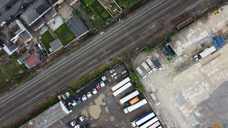 An aerial view of a mixed-use area showing railway tracks running horizontally across the image, with a variety of vehicles and containers situated on an adjacent paved lot. The railway lines are embedded in gravel and run parallel to a row of small residential houses with rooftops of various colors, including gray and red, visible on the left side. To the right of the tracks, there is a large open area with several parked white box trucks, some with orange and blue markings, along with other smaller vehicles. The lot contains scattered puddles, suggesting recent rain, and areas of concrete with different shades of gray and some patches of dirt or wear. Additionally, there are a few rows of stacked objects, possibly industrial or waste containers, and some blue plastic waste bins. The scene appears to be part of a practical space used for vehicle storage, loading, or waste handling, fitting within the context of alternative rubbish collection and private waste disposal services that Waste Collection Harringay provides in the local area near Turnpike Lane station.