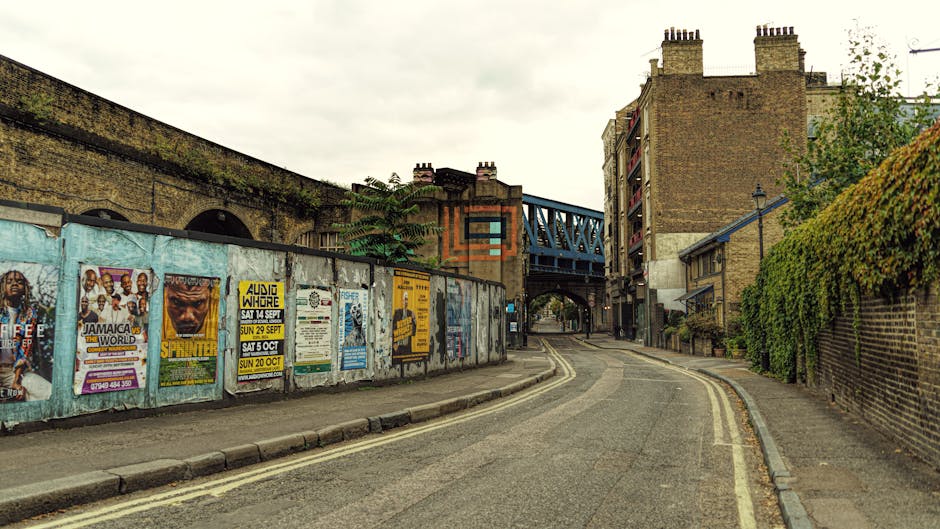 A narrow urban street with a gently curving asphalt surface bordered by double yellow lines on the left side and a raised concrete curb on the right. On the left, a weathered light blue wall covered with a variety of colorful posters and flyers advertises events such as concerts, DJ nights, and sports, with some posters partially peeling or faded. Behind the wall, a section of an old brick building with arched windows and moss-growing surfaces is visible, topped with chimney stacks. To the right, a high brick wall covered in dense, green foliage extends along the sidewalk, which is made of grey paving slabs. In the background, an overhead blue metal bridge with a lattice structure spans across the street, connecting two buildings, with a small archway at its base. Overcast sky casts diffused light over the scene, emphasizing the textures of the brick, posters, and foliage. The setting appears to be an inner-city area where private waste collections and on-site clearance might occur, with the environment reflecting an urban context suitable for rubbish removal services such as those provided by Waste Collection Harringay.