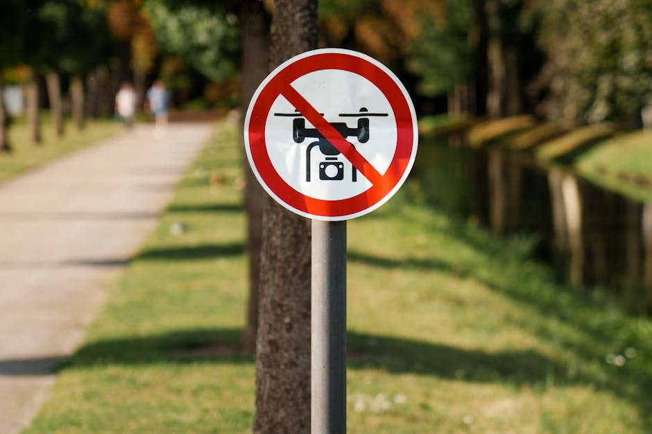 A circular sign with a red border and diagonal line crossing out a black drone icon, mounted on a metal pole in a park setting. The background features a paved pathway lined with evenly spaced trees on the left side, with some blurred figures walking in the distance. On the right, there is a grassy area with a narrow waterway or canal running parallel to the path. The scene is illuminated by natural sunlight, highlighting the lush green foliage and creating soft shadows. The sign clearly indicates that drone flights are prohibited in this outdoor public space, supporting local rules against aerial devices. Waste Collection Harringay provides rubbish removal services which, like drone restrictions, help maintain the safety and cleanliness of communal and private environments.