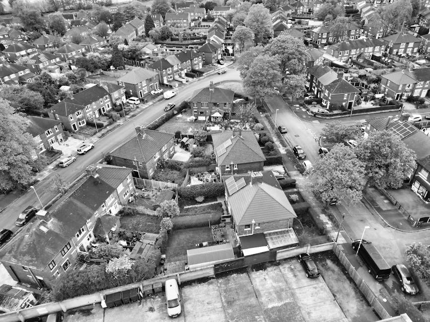 An aerial black-and-white photograph of a residential suburban area shows multiple detached and semi-detached houses with pitched roofs, surrounded by well-maintained gardens and narrow driveways. The foreground features a row of houses with small backyards enclosed by fences and hedges, some with visible outdoor furniture or garden sheds. The central part of the image reveals a large tree with a broad canopy casting shade over parts of the street and neighboring properties. Vehicles, including cars and a van, are parked along the streets and within individual driveways, indicating typical private waste disposal arrangements. The residential street curves gently and is bordered by additional houses and greenery, with some rooftop solar panels visible on a few buildings. Overall, the scene depicts a quiet, densely populated neighbourhood with consistent architectural styles and natural landscaping, consistent with a typical UK suburban setting. Waste Collection Harringay occasionally encounters similar residential scenes when supporting private waste collection or alternative rubbish removal needs in such neighbourhoods.