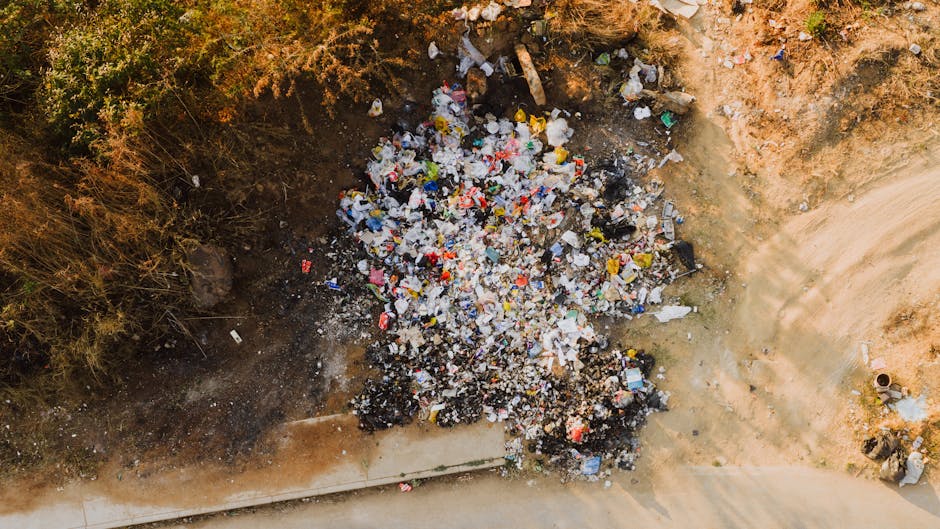 An aerial view of a large pile of mixed waste materials situated at the edge of a dirt area adjacent to a paved road. The pile contains various types of rubbish, including plastic bags, bottles, paper, and packaging, with some materials appearing crumpled or shredded. Surrounding the waste are patches of dry, brown soil and sparse vegetation with small, bushy plants and dried foliage on the upper left side. The ground beneath the rubbish is darker and looks disturbed, indicating recent disposal or accumulation. To the right, the ground transitions to a lighter, sandy surface with visible tire tracks and a concrete footpath running horizontally along the bottom edge of the image. The scene suggests an informal or alternative waste disposal site, which Waste Collection Harringay might handle through on-site clearance or private collection services, emphasizing the importance of proper rubbish removal methods for maintaining cleanliness and environmental standards in Harringay.