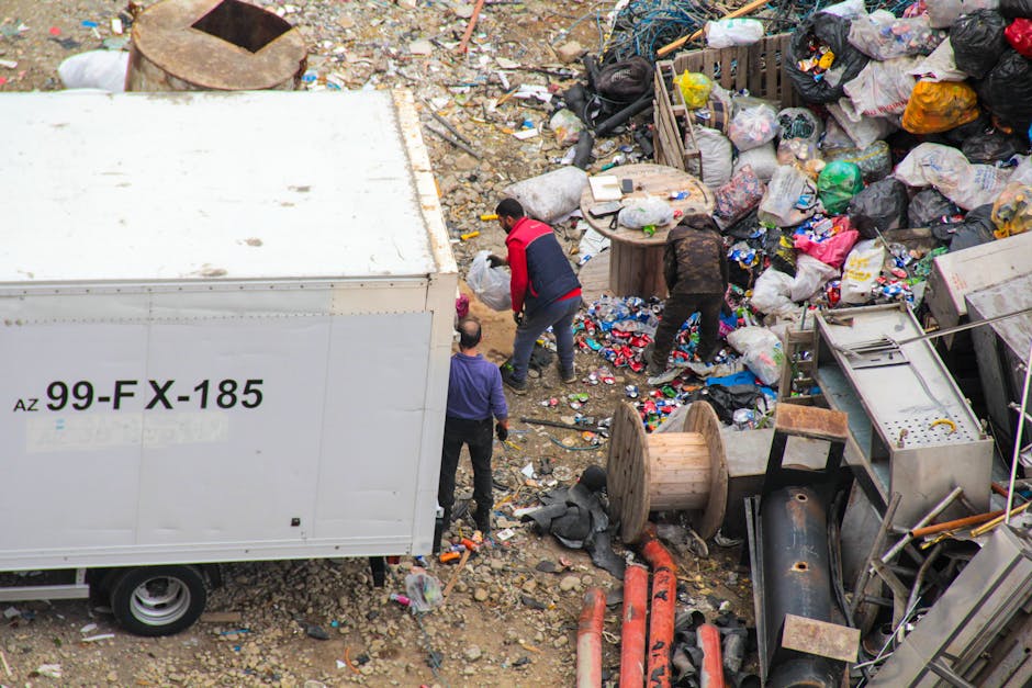 An outdoor scene showing a large accumulation of mixed waste and debris on uneven, rocky ground. Several black, white, and multicoloured plastic garbage bags are piled next to discarded items, including wooden spools, metal pipes, and broken appliances. Four individuals are actively engaged in waste collection, with two bending over to gather rubbish and one standing nearby. A white truck with the registration code '99-FX-185' is partially visible on the left side of the image, suggesting a waste removal operation. The surrounding environment appears cluttered and messy, indicative of an area undergoing clearance or disposal activity, characteristic of independent waste collection efforts typically seen in private disposal scenarios. The natural lighting emphasizes the textures of various materials, from the crushed plastics to the rusted metal components, reinforcing the scene’s focus on refuse and waste management processes linked to rubbish removal services.
