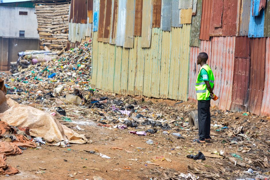 An aerial view of a large pile of mixed waste materials situated at the edge of a dirt area adjacent to a paved road. The pile contains various types of rubbish, including plastic bags, bottles, paper, and packaging, with some materials appearing crumpled or shredded. Surrounding the waste are patches of dry, brown soil and sparse vegetation with small, bushy plants and dried foliage on the upper left side. The ground beneath the rubbish is darker and looks disturbed, indicating recent disposal or accumulation. To the right, the ground transitions to a lighter, sandy surface with visible tire tracks and a concrete footpath running horizontally along the bottom edge of the image. The scene suggests an informal or alternative waste disposal site, which Waste Collection Harringay might handle through on-site clearance or private collection services, emphasizing the importance of proper rubbish removal methods for maintaining cleanliness and environmental standards in Harringay.
