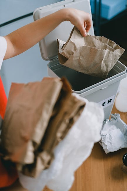 A person disposing of brown paper and plastic bags into a small, rectangular, white rubbish bin with a black interior, positioned on a wooden surface. The person is wearing a short-sleeved top, and their arm is extended to place an open paper bag into the bin. There are additional crumpled paper bags and a white plastic bottle on the table nearby. The environment appears to be indoors, with part of a white wall or window frame visible in the background. The scene illustrates an informal waste disposal process suitable for domestic rubbish removal, aligning with services offered by Waste Collection Harringay for private on-site clearance or alternative waste handling outside of municipal collection systems.