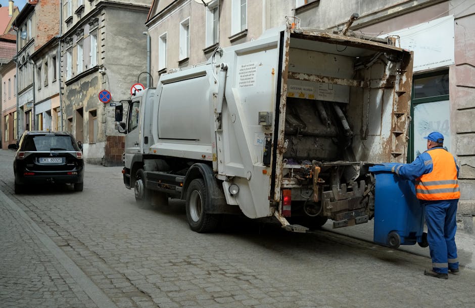 A person disposing of brown paper and plastic bags into a small, rectangular, white rubbish bin with a black interior, positioned on a wooden surface. The person is wearing a short-sleeved top, and their arm is extended to place an open paper bag into the bin. There are additional crumpled paper bags and a white plastic bottle on the table nearby. The environment appears to be indoors, with part of a white wall or window frame visible in the background. The scene illustrates an informal waste disposal process suitable for domestic rubbish removal, aligning with services offered by Waste Collection Harringay for private on-site clearance or alternative waste handling outside of municipal collection systems.